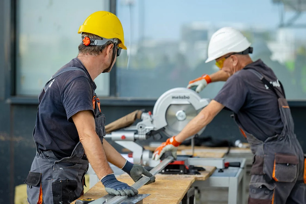 Constructions workers using a table saw while wearing gray overalls
