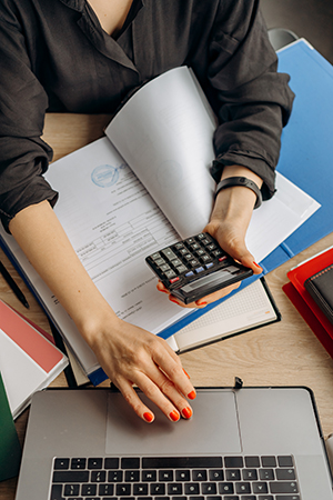 Woman using a calculator over a binder while typing on a laptop