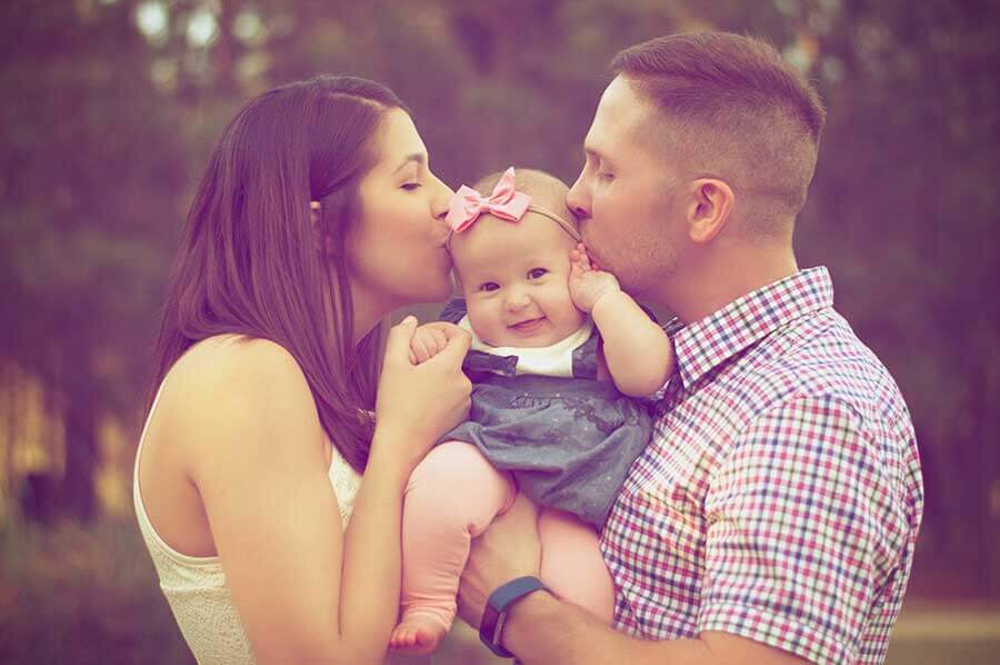 Man and woman kissing a baby with a pink bow