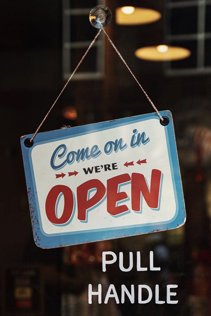 White and blue sign hanging from glass that says come in we're open