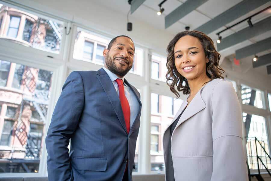 Two people smiling in front of a window overlooking the city