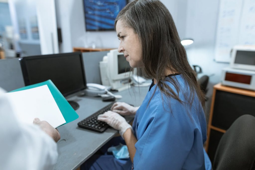 Woman in blue scrubs looking over notes while typing on a keyboard