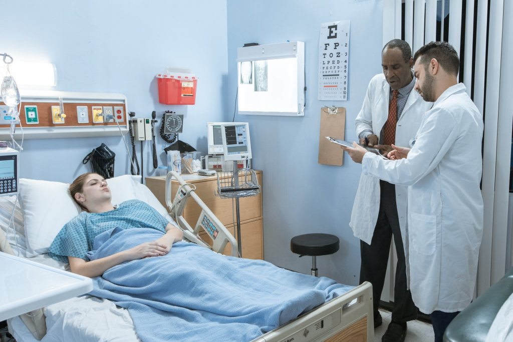 Two doctors looking over a patient file in a hospital room while a patient sleeps