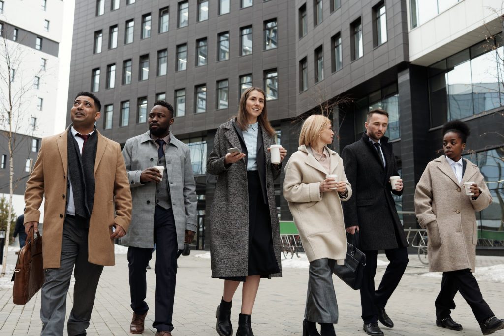 People walking down a sidewalk behind a large brown building wearing trench coats